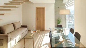 A modern living room with a sofa, a glass dining table, and chairs. There is a wooden door and a window with a view of buildings. - Beautiful Penthouse Duplex @ the best area in La Paz (Zona Calacoto La Paz)