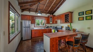 A kitchen with wooden cabinets, a stainless steel refrigerator, a large island with bar stools, and a window overlooking greenery. - Exclusive Full House near Beaches & Downtown (Roatan)
