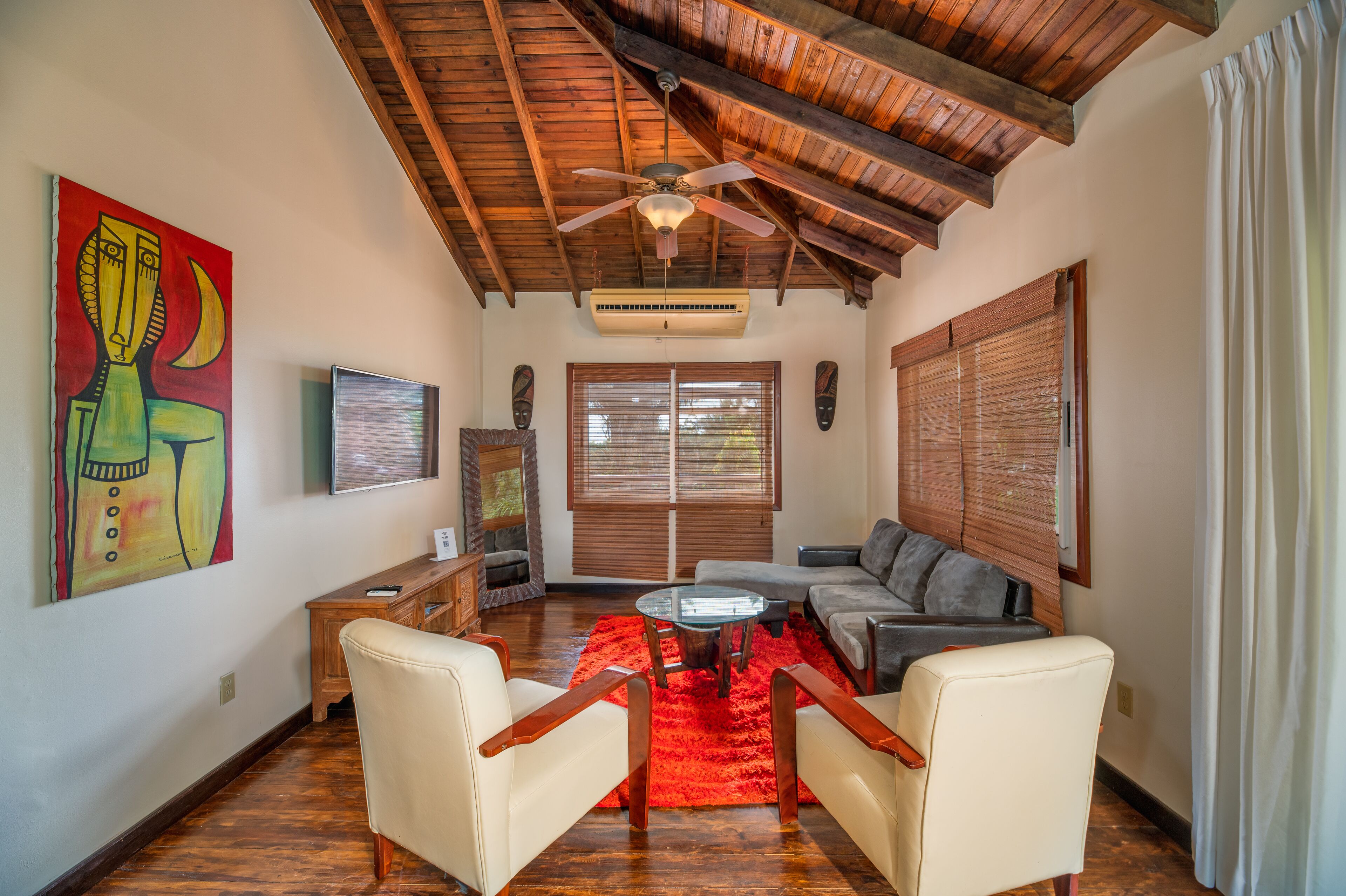 A living room with a wooden ceiling, a red rug, a sofa, two armchairs, a TV, and a painting on the wall.