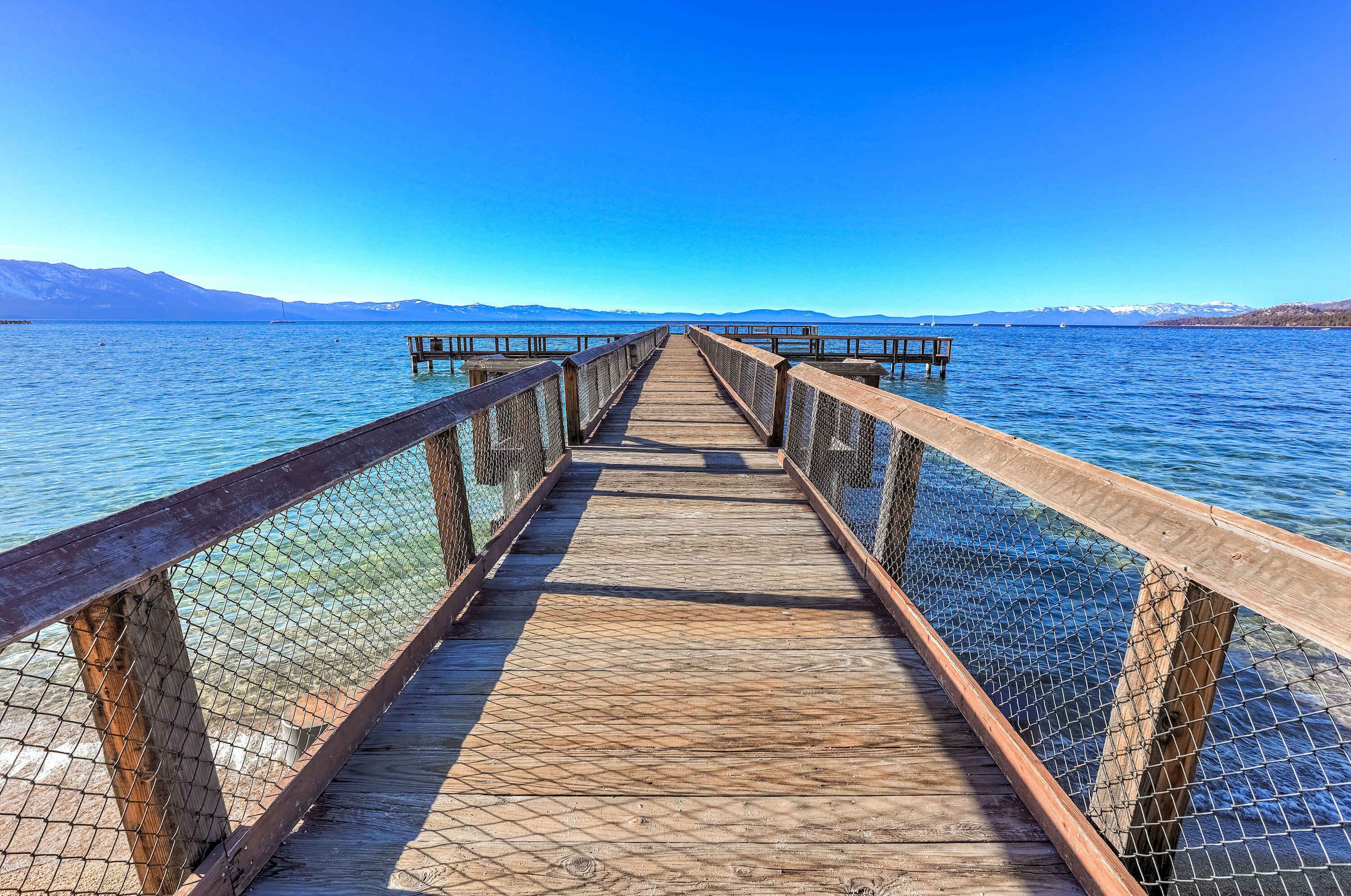 A wooden pier extending into a calm lake with clear blue water and distant mountains.