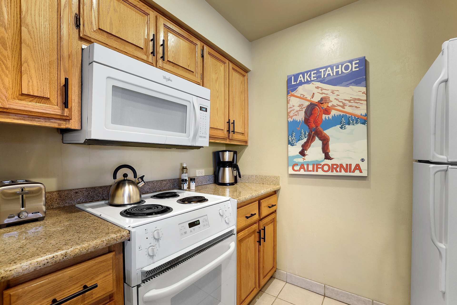 Una cocina con armarios de madera, una estufa blanca, un microondas y un hervidor. Hay una cafetera y un tostador en la encimera. En la pared, un cartel de un hombre pescando en el Lago Tahoe, California.