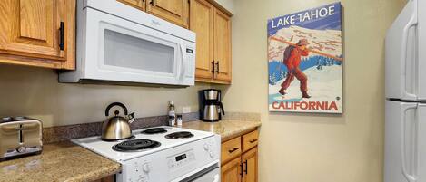 Una cocina con armarios de madera, una estufa blanca, un microondas y un hervidor. Hay una cafetera y un tostador en la encimera. En la pared, un cartel de un hombre pescando en el Lago Tahoe, California.
