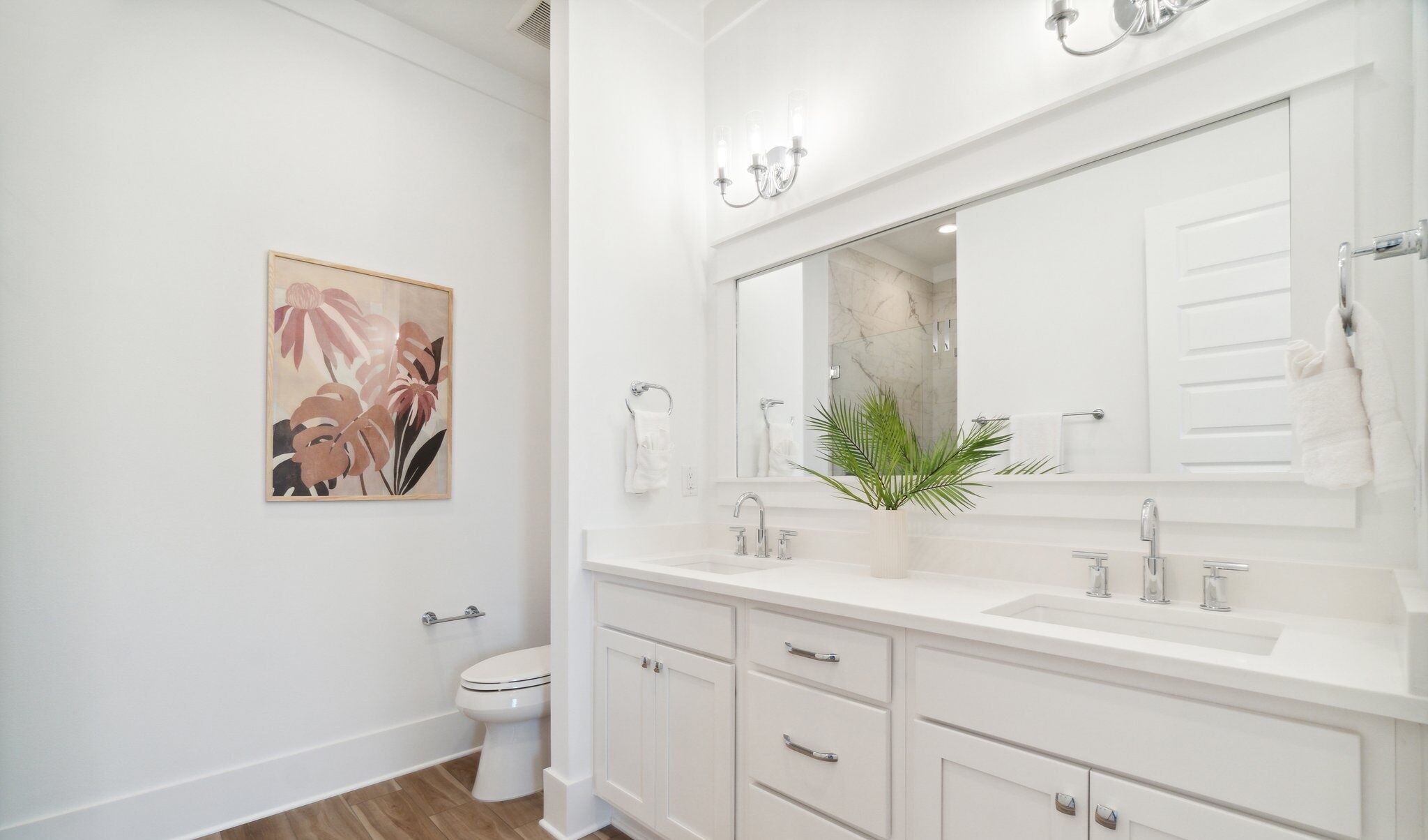 Une salle de bain moderne avec un lavabo, un meuble-lavabo et des toilettes, le tout de couleur blanche. Un grand miroir est placé au-dessus du lavabo, et un tableau représentant des motifs floraux est accroché au mur.