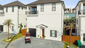 A modern two-story residential building with a garage, a covered driveway, and a small front yard with palm trees. - ✨ Summer Lane | Golf Cart | Heated Private Pool*  & Walk to Beach | Elevator | FREE Parasailing (Santa Rosa Beach)