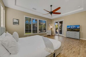 Una camera da letto con un letto, una ventola a soffitto, una TV e vista sull'esterno attraverso ampie finestre.