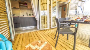 Un patio descubierto con una mesa de comedor y sillas, una pequeña mesa redonda con dominó, y un área de cocina visible a través de puertas correderas de vidrio.