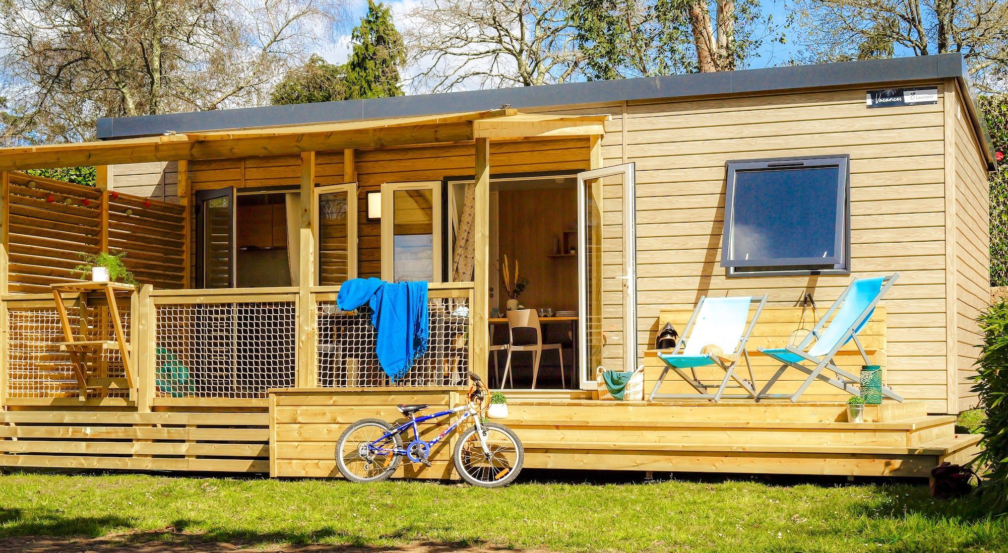 Een houten chalet met een overdekte veranda, een fiets en twee loungestoelen.