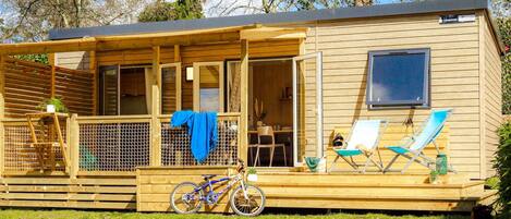 Een houten chalet met een overdekte veranda, een fiets en twee loungestoelen.