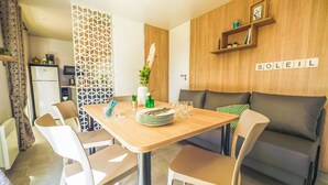 Un espace repas moderne avec une table en bois, des chaises blanches et un canapé. On y trouve un panneau mural décoratif et une étagère avec de petits objets.