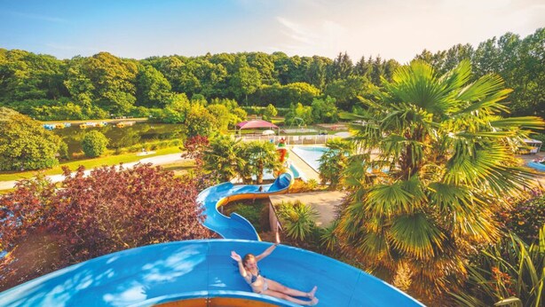 Un parc aquatique avec une personne sur un toboggan bleu, entourée de verdure et d’une zone de piscine.