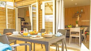 Une table de petit-déjeuner dressée avec divers aliments et une tasse de café, sur une terrasse en bois donnant sur la cuisine.