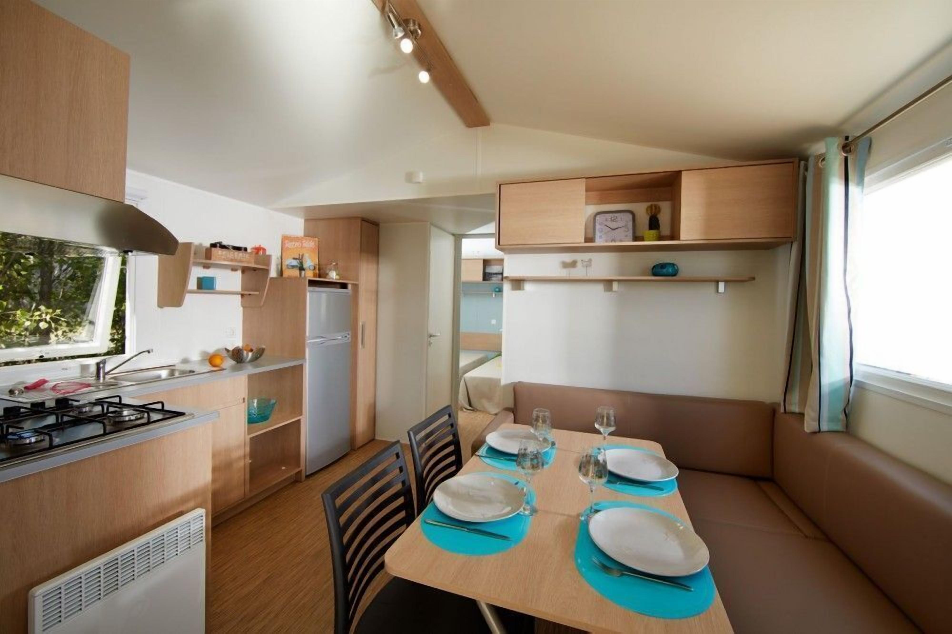 A compact kitchen with a gas stove, sink, and a refrigerator. There is a dining area with a table set for four, chairs, and a bench.