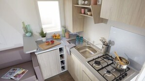 Cocina compacta con estufa a gas, lavamanos y una pequeña encimera con tabla de cortar y frutas.