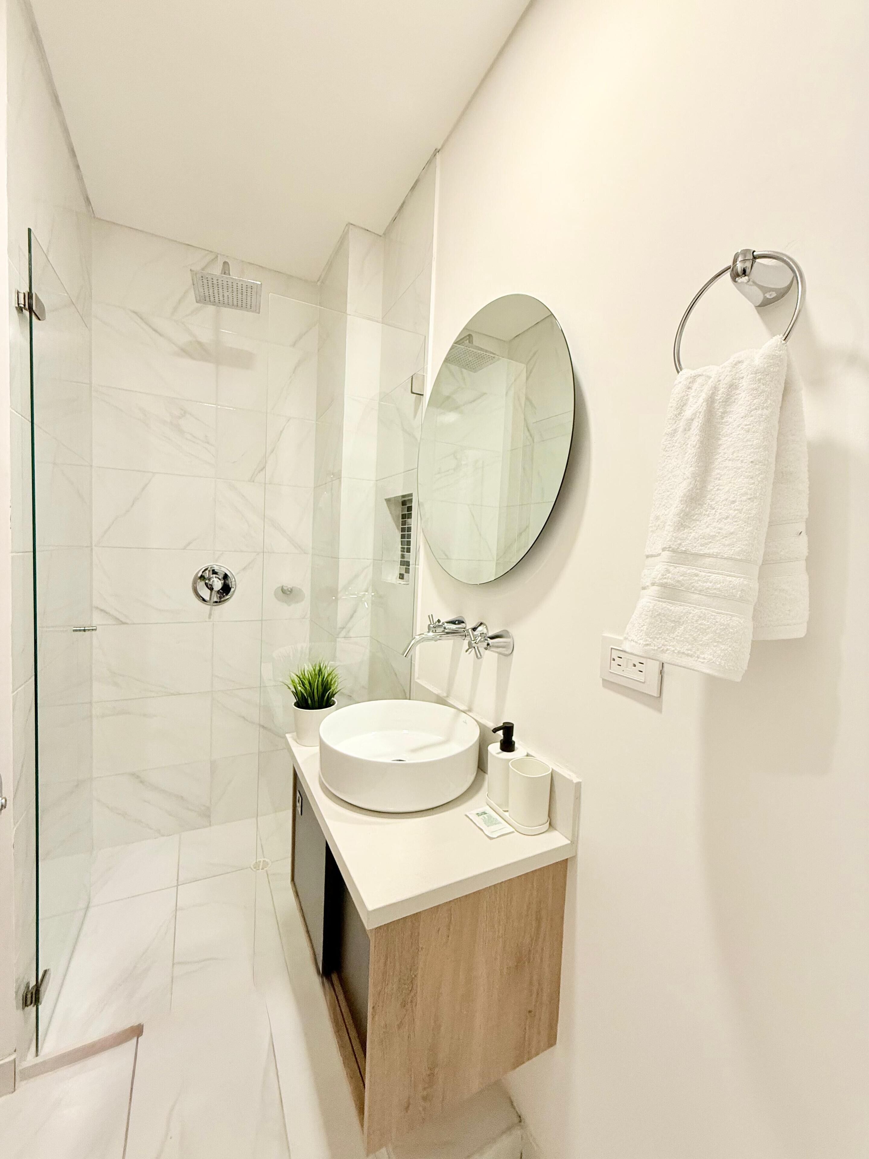 Un baño moderno con cabina de ducha de vidrio, espejo redondo, lavabo con tocador de madera y un perchero para toallas.
