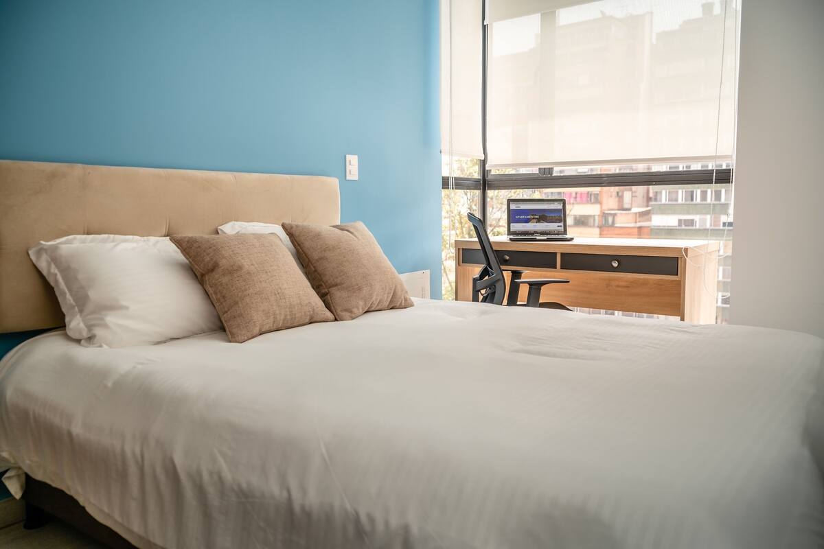 A bedroom with a bed, two pillows, a desk with a laptop, and a window with a city view.
