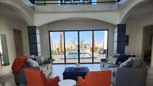Ein geräumiges Wohnzimmer mit zwei Sofas, einem Couchtisch und orangefarbenen Sesseln. Große Schiebegläser führen zu einem Außenpoolbereich.