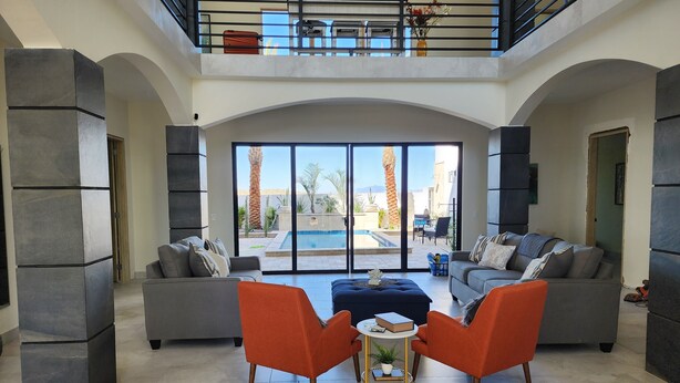 Ein geräumiger Wohnbereich mit Blick auf einen Pool, grauem Sofa, orangefarbenen Sesseln und einem kleinen Tisch mit einer Pflanze.