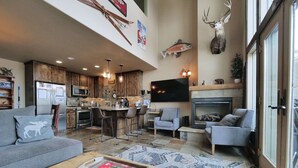 A modern living room with a grey sofa, a wooden coffee table with a magazine, a fireplace, and a kitchen area with wooden cabinets and a TV mounted on the wall. - Kirkwood Mountainside Cabin w/Views, Walk to Lifts (Kirkwood)