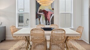 Un coin repas comprenant une table rectangulaire, des chaises en osier, un luminaire suspendu moderne et un grand tableau abstrait accroché au mur.
