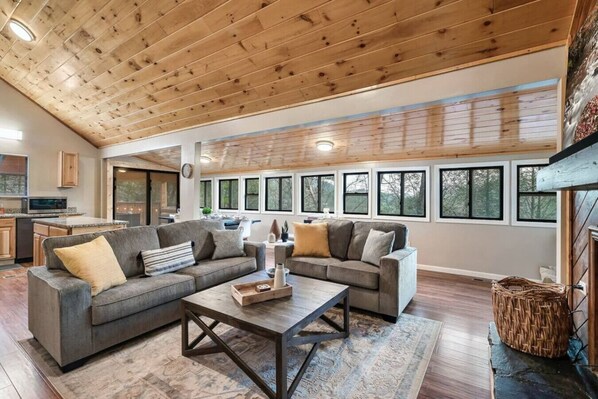 A spacious living area with a wooden ceiling, large windows, and a kitchen in the background. - Smokey Acres Getaway (Sevierville)