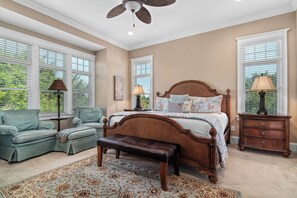 Quarto com cama, duas cadeiras, um banco, mesa de cabeceira e ventilador de teto.