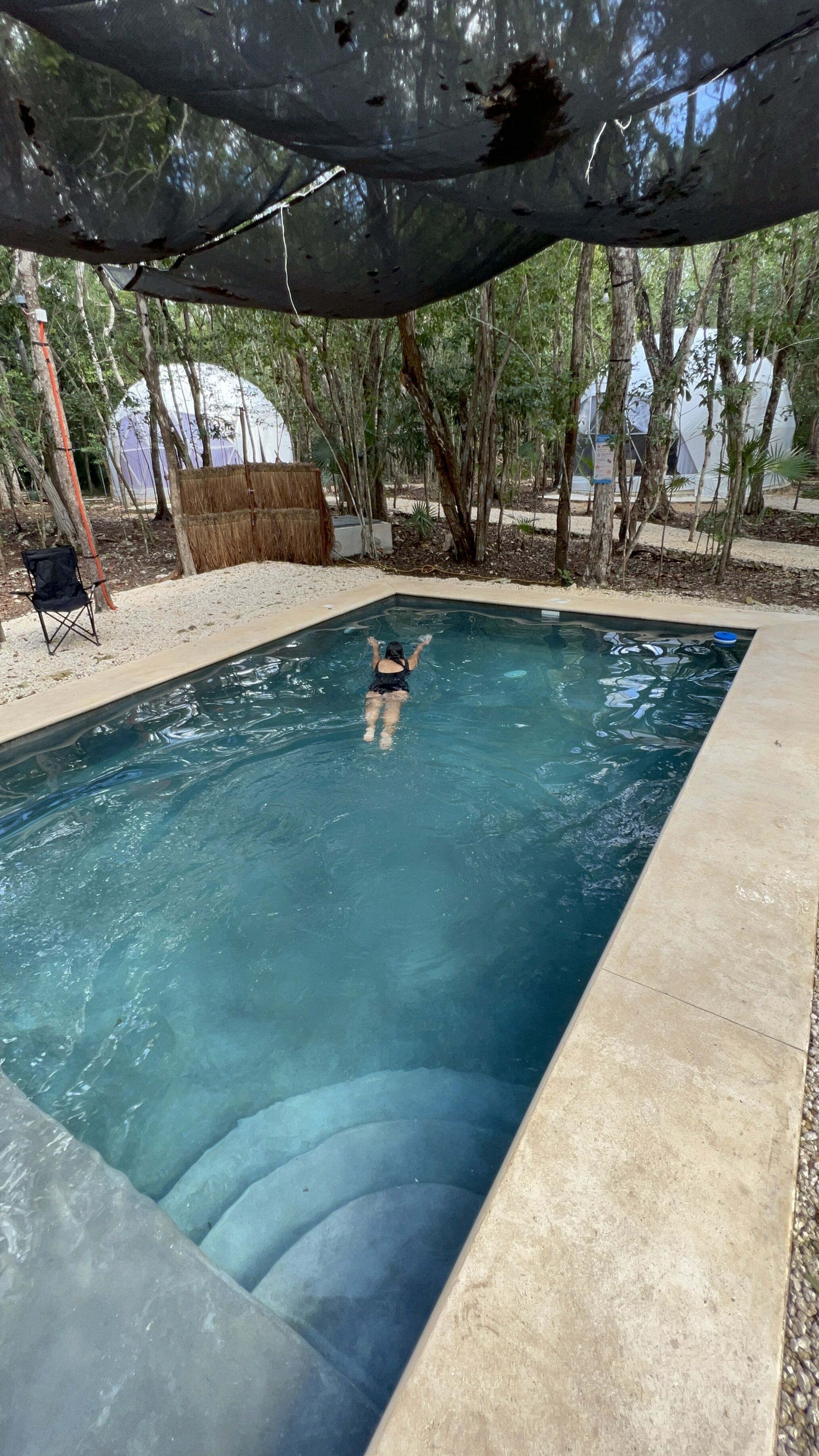 A swimming pool with a person swimming, surrounded by trees and a covered area.
