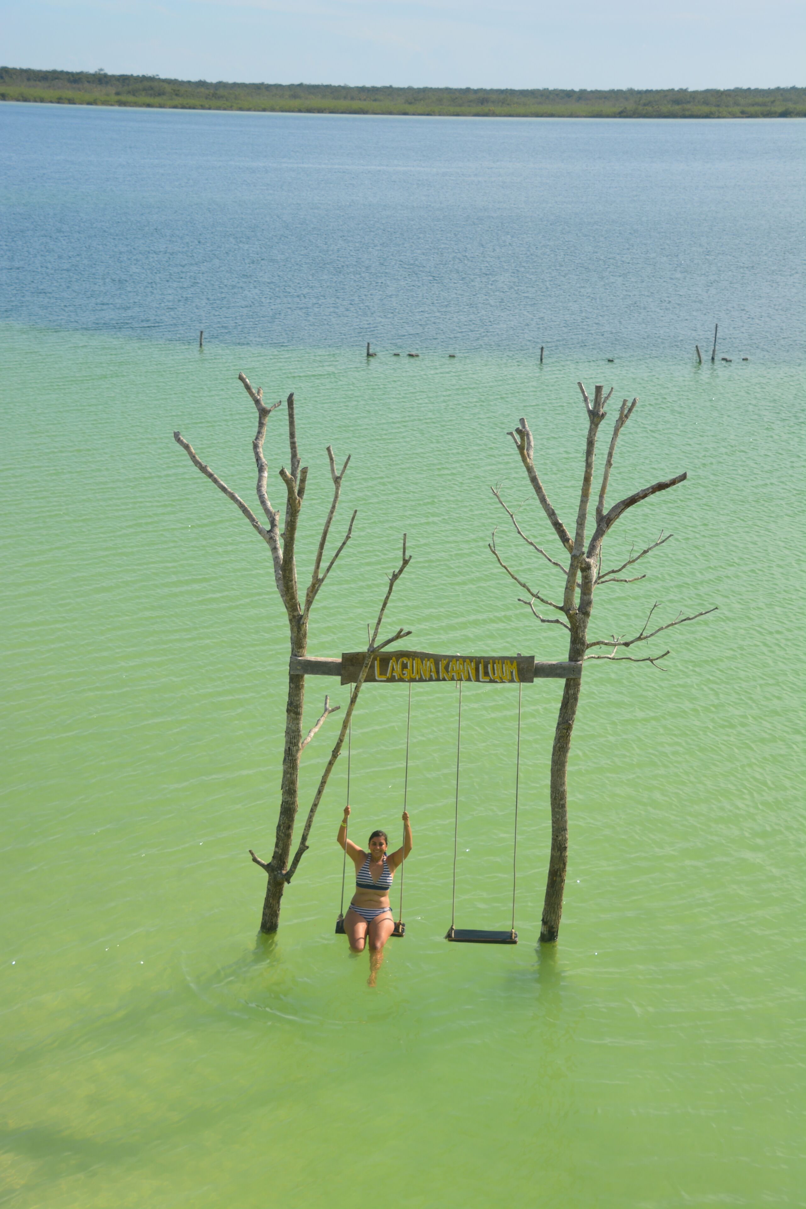 Une personne est assise sur une balançoire au-dessus d’une étendue d’eau, et un panneau indique : « LAGUNA KANN LUHM ».