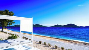 A beachside view with a clear sky, a sandy beach, and a white structure with a view of the sea and distant hills.