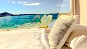 Een bed met uitzicht op een heldere blauwe zee en verre bergen door een raam.