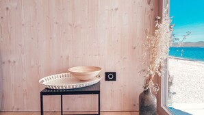 Een kamer met houten vloer, een klein zwart metalen tafeltje met een geweven kom, een vaas met gedroogde planten, en uitzicht op een strand door een groot raam.