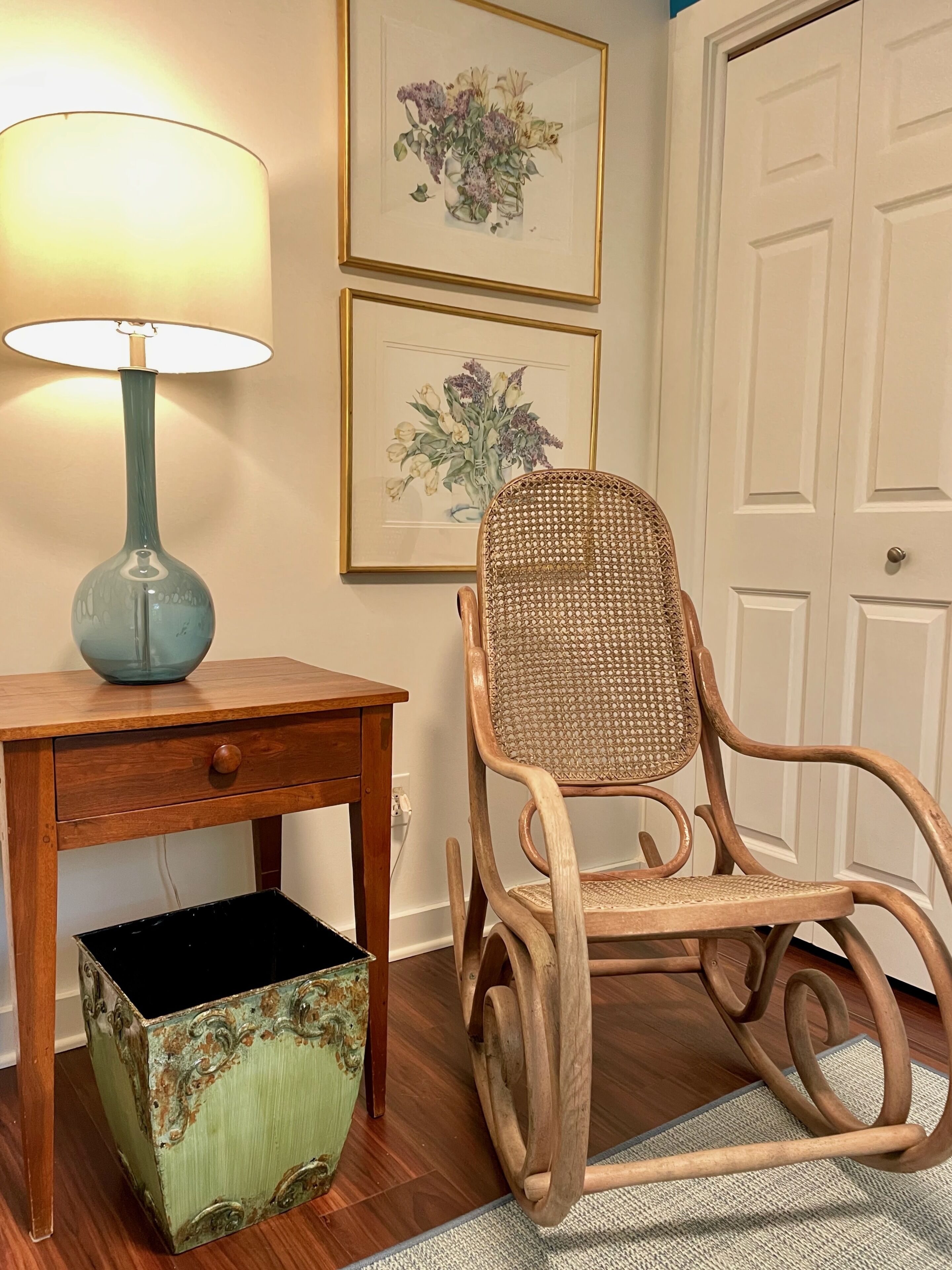 A wooden chair with a woven backrest, a small wooden table with a drawer, a lamp, and a trash bin.