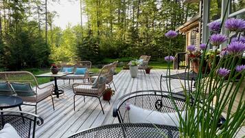 Une terrasse en bois, agréée de mobilier d’extérieur, entourée d’arbres et bordée d’un bâtiment doté de grandes fenêtres.