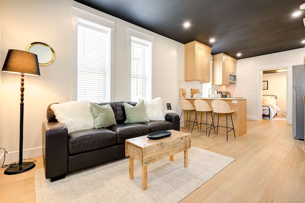 Een moderne woonkamer met een donker leren bank, een houten salontafel en een vloerlamp. Er is een keuken met houten kasten en een barkruk.