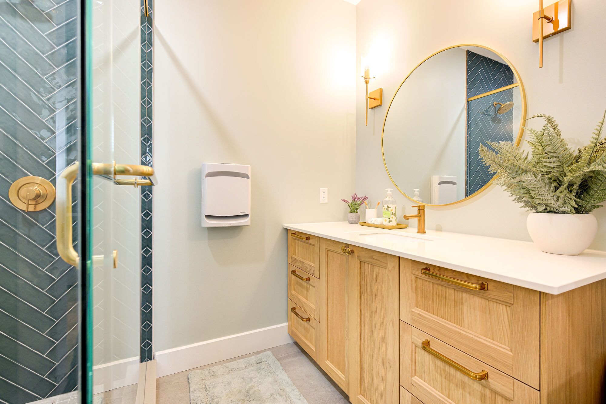 A bathroom with a glass shower door, a white sink with a gold faucet, a wooden vanity with drawers, a round mirror, and a potted plant.