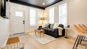 A living room with a black leather sofa, a wooden coffee table, a wicker chair, and a small round table with a lamp.