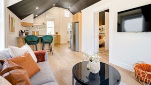 A modern living room with a sofa, a coffee table, and a TV. There is a kitchen in the background with a refrigerator and a dining area with chairs. - The Willow ⍋ | 1 Bed 1 Bath | 2nd Storey Suite (Kitchener)