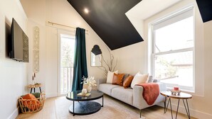 A modern living room with a sofa, a coffee table, and a small side table. There is a large window providing natural light and a view of the outside. - The Willow ⍋ | 1 Bed 1 Bath | 2nd Storey Suite (Kitchener)
