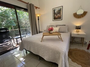 A bedroom with a bed, a bedside table with a lamp, a framed picture on the wall, and a view of the outdoors through a window.