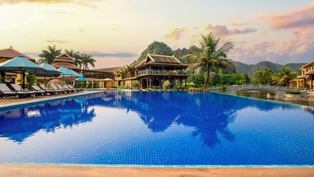 A swimming pool with lounge chairs and umbrellas, surrounded by buildings and palm trees. - Emeralda Resort Tam Coc (Nam Hoa Lu)