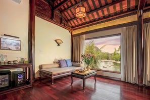 A room with a sofa, a table with a vase, a painting, and a view of a body of water. - Emeralda Resort Tam Coc (Nam Hoa Lu)