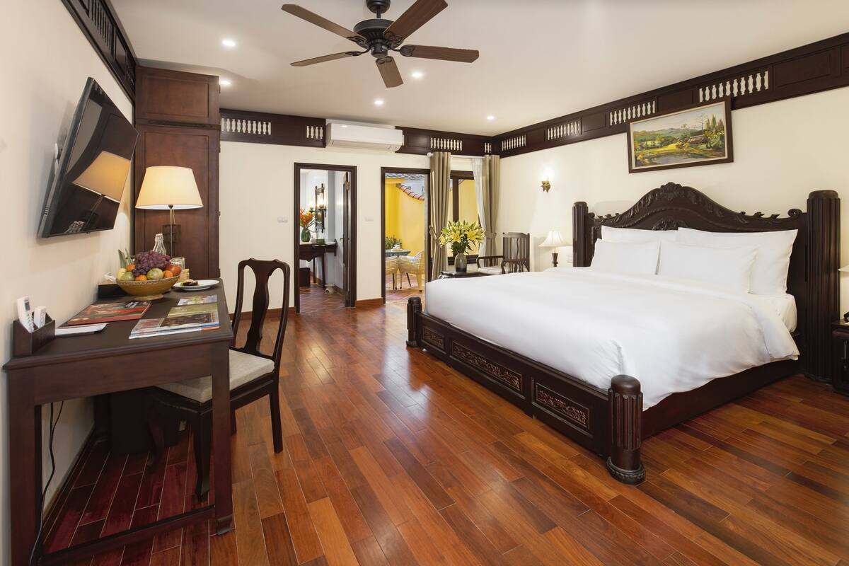 A hotel room with a large bed, wooden flooring, a desk with a lamp, a television, and a ceiling fan.