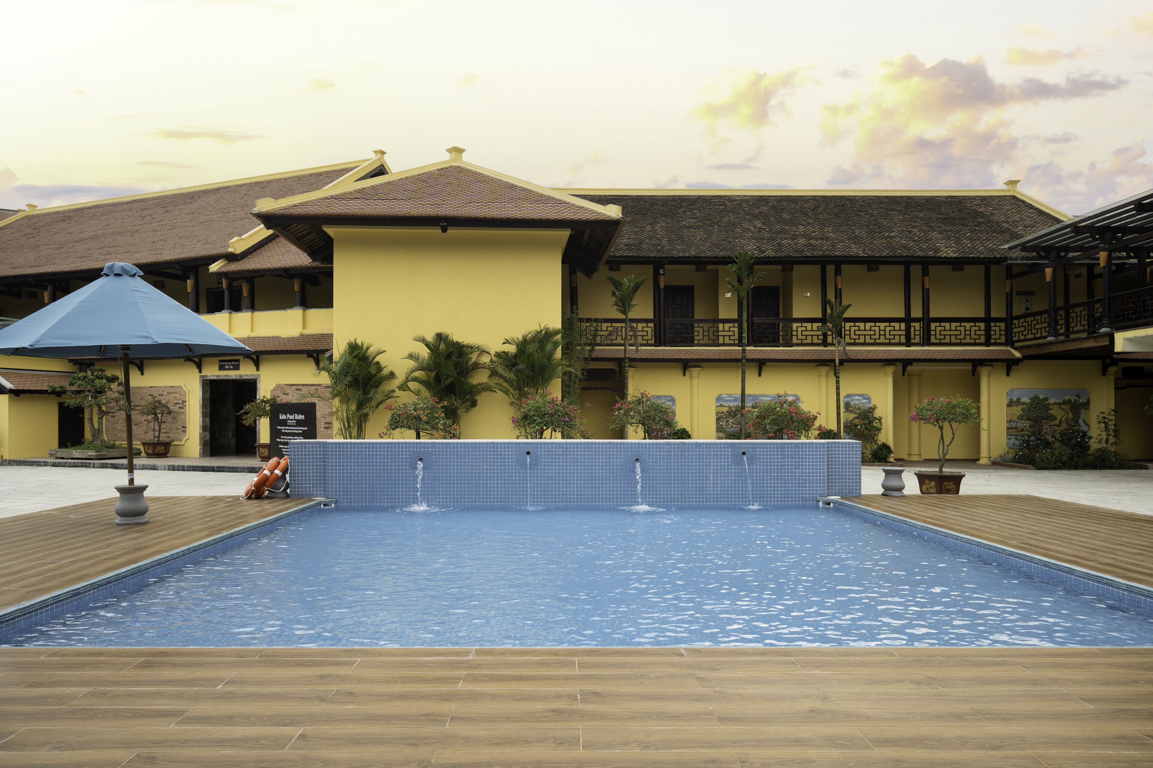 A swimming pool with a wooden deck, surrounded by a yellow building with a balcony.