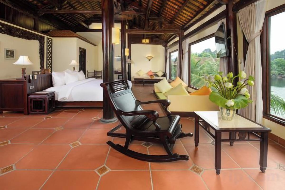 A hotel room with a bed, a rocking chair, a table with a vase of flowers, and a view of the outdoors.