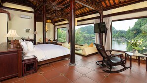 A spacious room with a large bed, a rocking chair, a desk, and a view of a lake. - Emeralda Resort Tam Coc (Nam Hoa Lu)