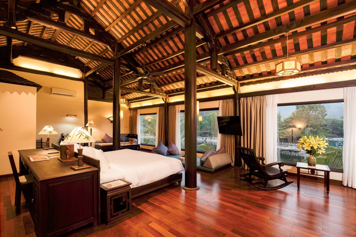 A hotel room with a wooden ceiling, a bed, a desk with a lamp, a chair, and a view of the outdoors.
