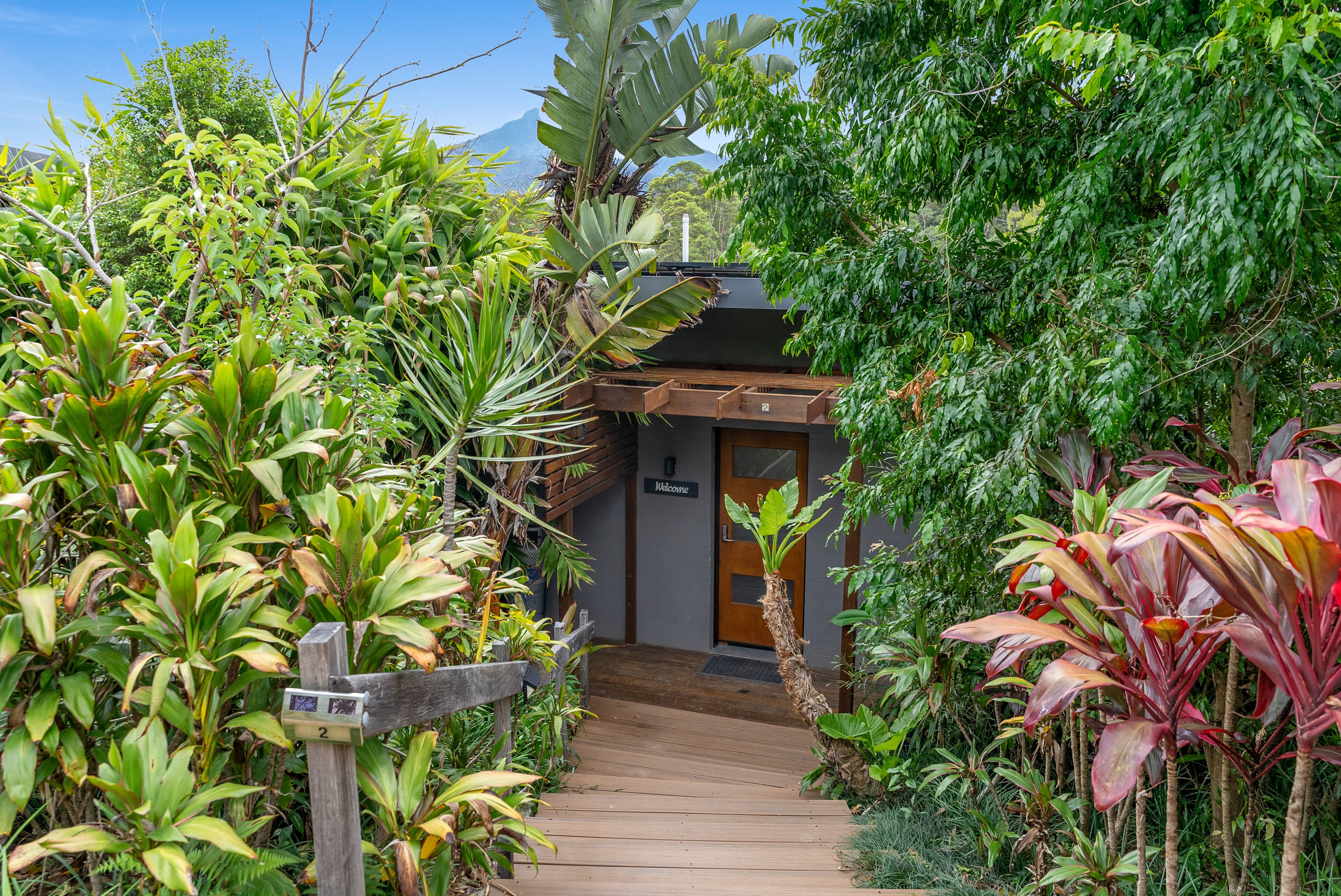 Un sendero de madera que conduce a un edificio con un letrero que dice 'Welcome', rodeado de exuberante follaje y plantas tropicales.