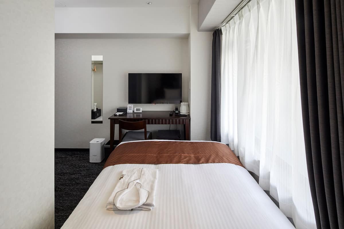 A hotel room with a bed, a desk, a flat-screen TV, and a window with curtains.