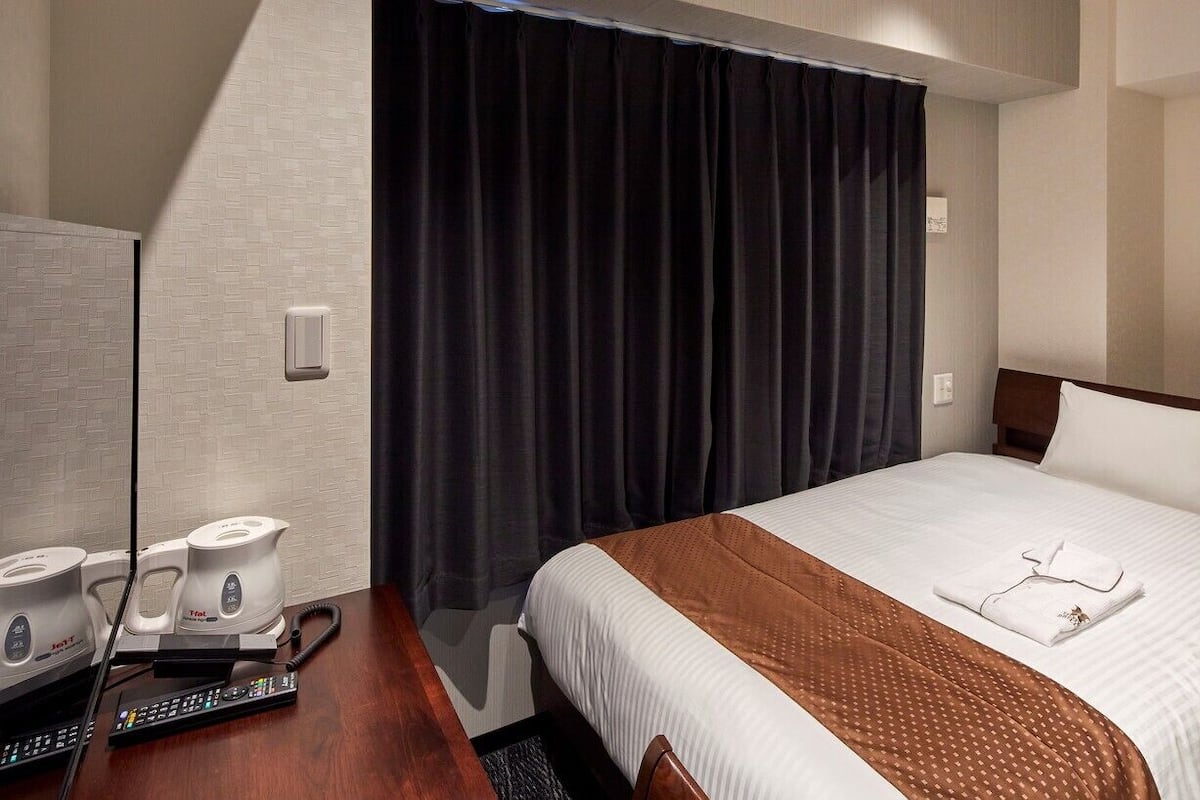 A hotel room with a bed, a desk with a telephone and a coffee maker, a dark curtain, and a painting on the wall.