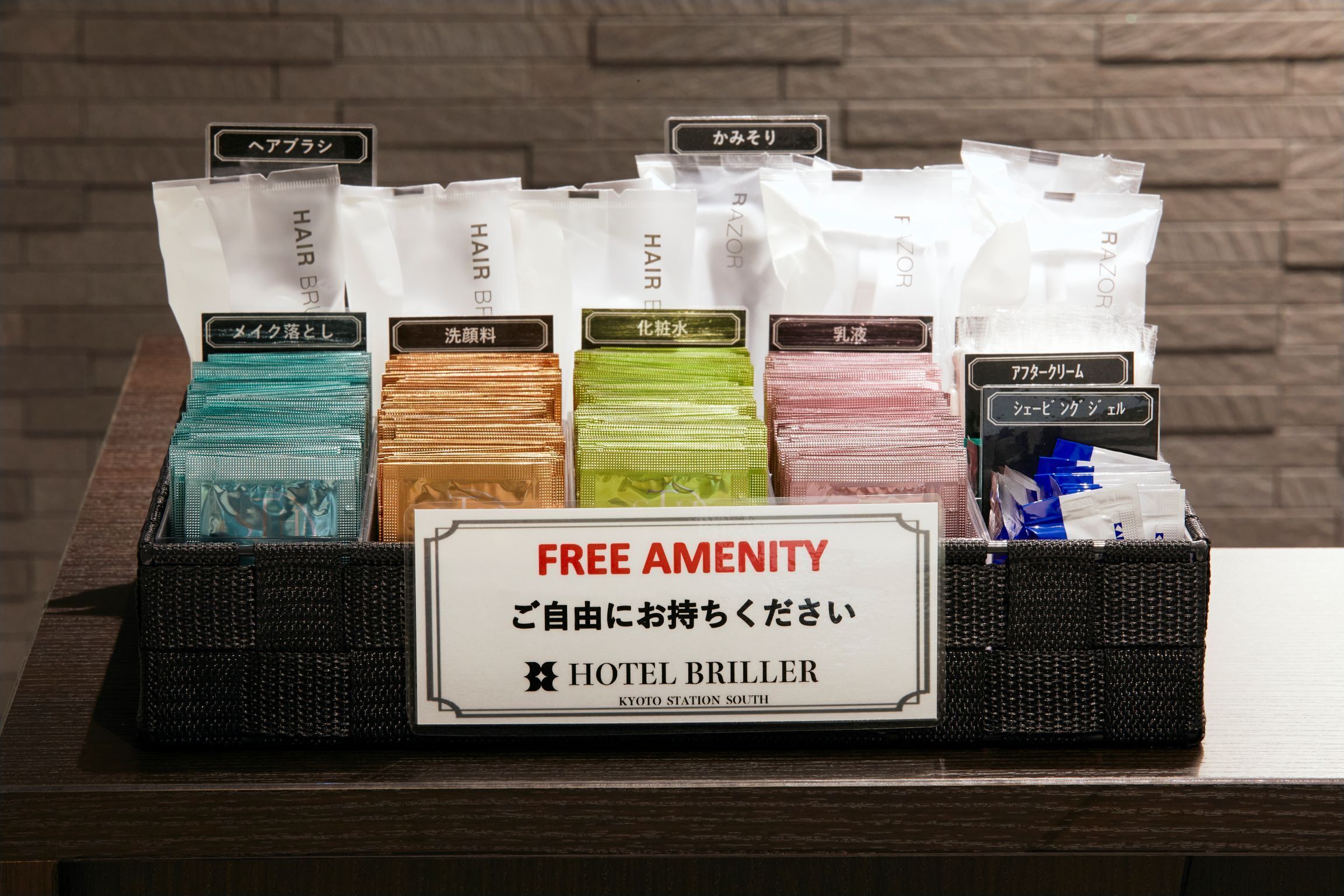 A display of hair care products with labels in Japanese, placed on a table with a sign reading 'FREE AMENITY' and 'HOTEL BRILLER KYOTO STATION SOUTH'.