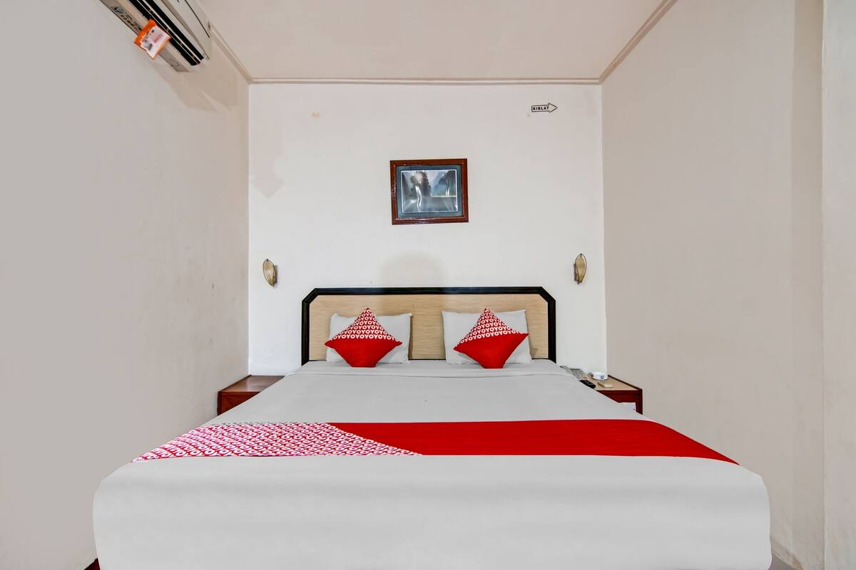 Un lit bien fait, recouvert d’une couvre-lit rouge et blanche, avec deux oreillers rouges et une petite table de chevet surmontée d’une lampe.